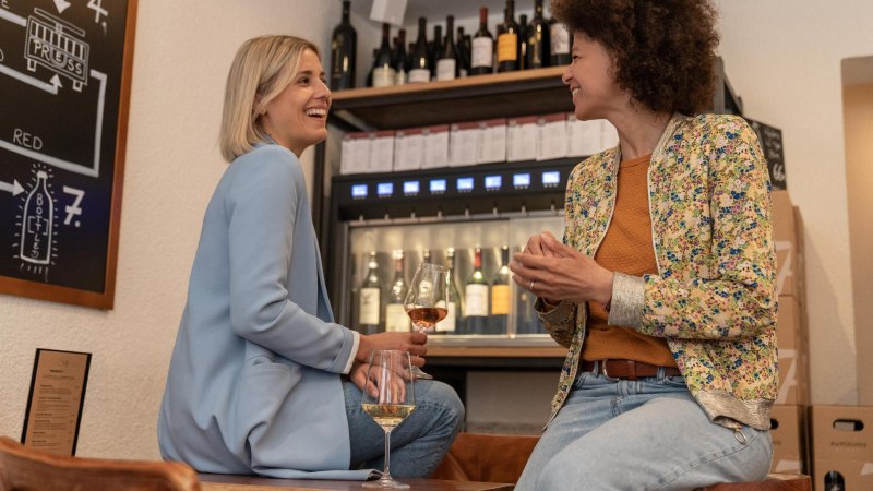 Zwei Frauen sitzen auf hohen Stühlen und lachen einander zu. Eine hält in der Hand ein Weinglas. Im Hintergrund ein Weinspender mit zapfbaren Weinflaschen., © Stuttgart-Marketing GmbH, Martina Denker