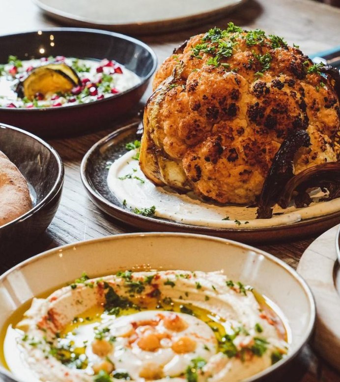 Ein Tisch mit verschiedenen orientalischen Gerichten, darunter gerösteter Blumenkohl, Hummus mit Kichererbsen, Pita-Brot und Shakshuka mit pochierten Eiern in Tomatensauce., © yafa, Stuttgart