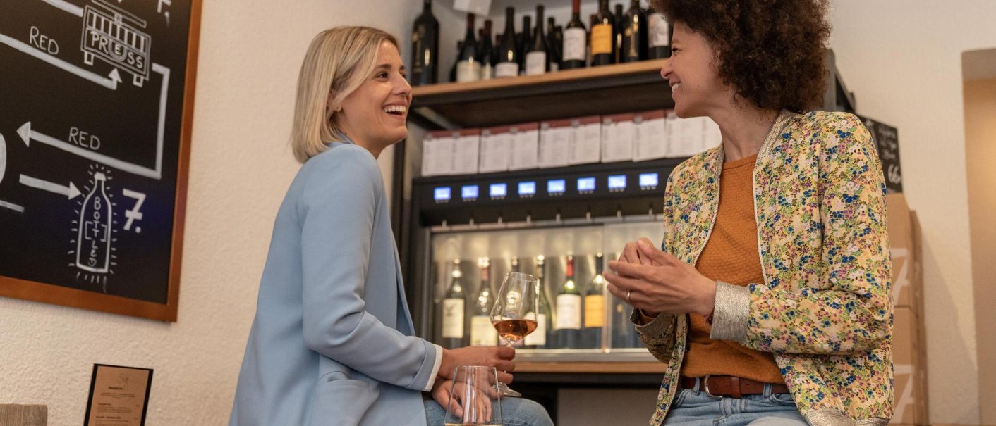 Two women are sitting on high stools, one of them is holding a glas of wine. They are laughing at each other. In the background is a wine dispenser with wine bottles that can be tapped., © Stuttgart-Marketing GmbH, Martina Denker