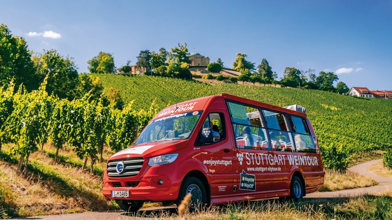 Eine Luftaufnahme zeigt den roten Weintour-Bus der Stuttgarter Citytour, wie er auf einer asphaltierten Straße durch einen Weinberg fährt. Das Laub des Weins ist saftig grün und die Szene ist lichtdurchflutet. , © SMG, Thomas Niedermüller 