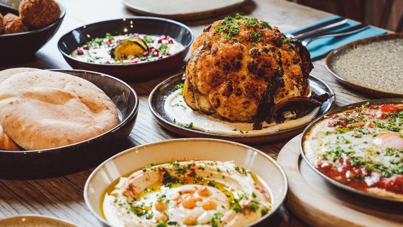 Ein Tisch mit verschiedenen orientalischen Gerichten, darunter gerösteter Blumenkohl, Hummus mit Kichererbsen, Pita-Brot und Shakshuka mit pochierten Eiern in Tomatensauce., © yafa, Stuttgart