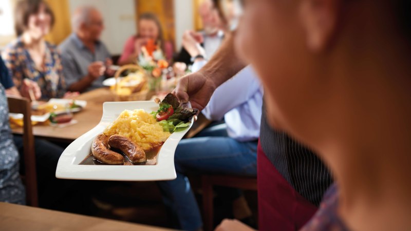 Ein Teller mit geschmorten Bratwürsten, Kartoffelpüree und Salat wird in einem gemütlichen Restaurant serviert. Im Hintergrund sitzen Menschen an einem Holztisch und genießen ihr Essen in geselliger Runde., © SMG, Jean-Claude Winkler