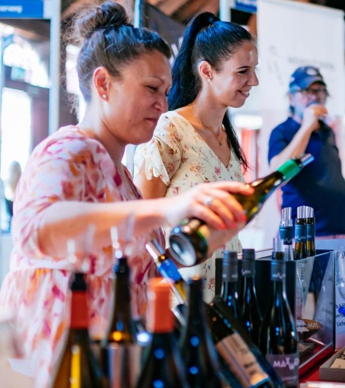 Two women serve wine at a tasting event, surrounded by wine bottles. In the background there are some guests tasting wine at a different table., © Stuttgart-Marketing GmbH, Thomas Niedermüller