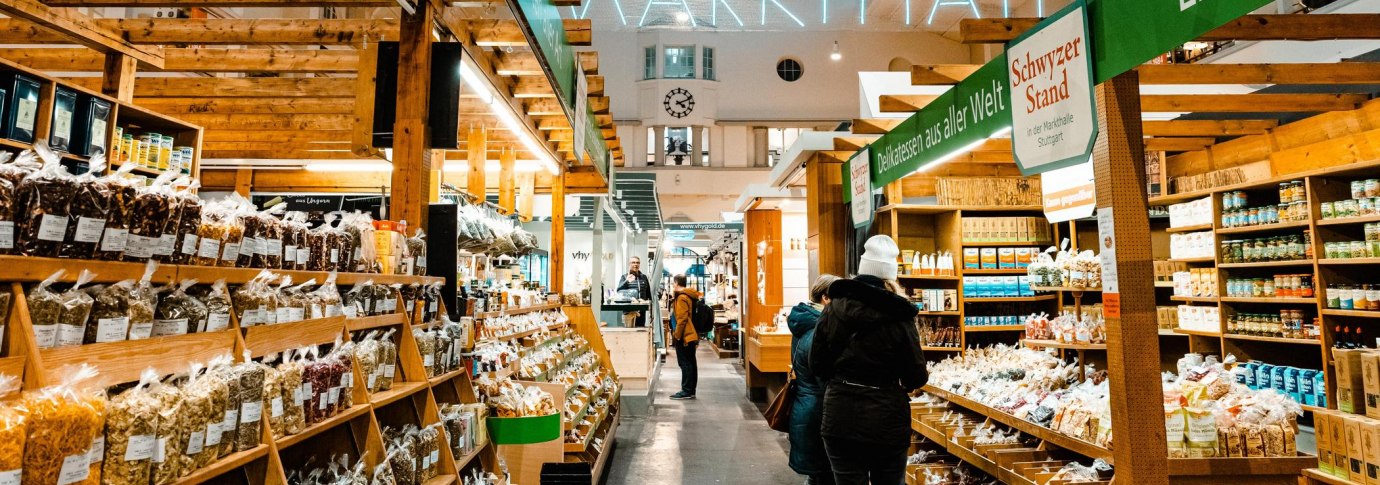 Bild aus dem Inneren der Markthalle. Gezeigt wird ein Gang, an dem es links und rechts Gewürze zu kaufen gibt. Zwei Personen in warmer Kleidung betrachten die Auslage auf der rechten Seite., © Stuttgart-Marketing GmbH, Sarah Schmid